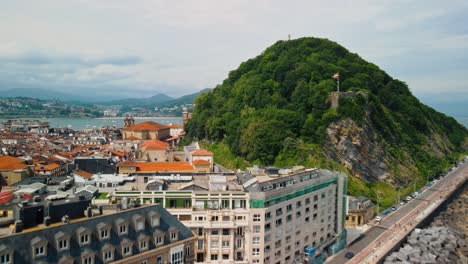 Vibrant-orange-building-facade-and-lush-vegetated-coastal-hillside-of-San-Sebastian-in-Spain,-city-establishing-reveal-of-lagoon