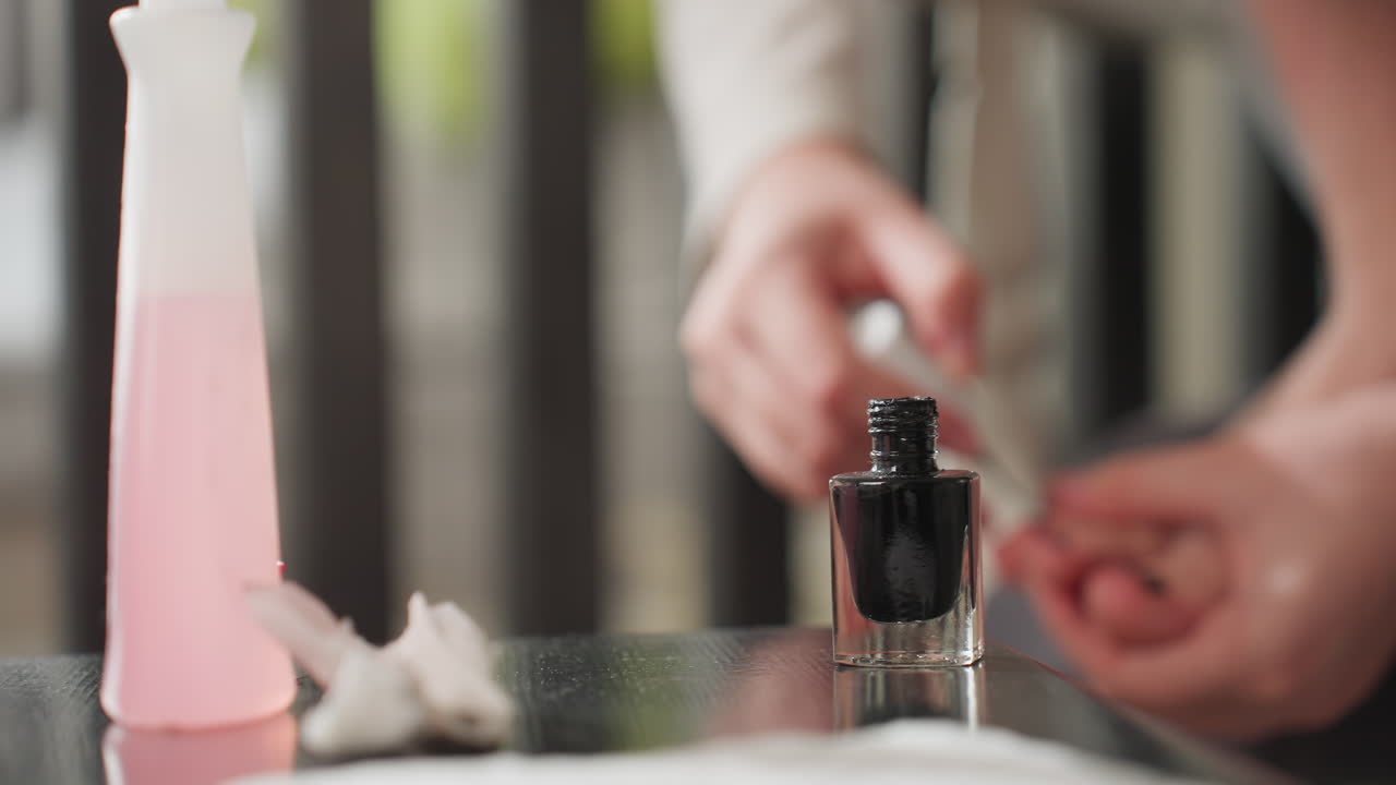 Round view shows black nail polish bottle on table beside pink remover with cap off, blurred fair skinned girl in background carefully applying color to toes, glossy surface reflections