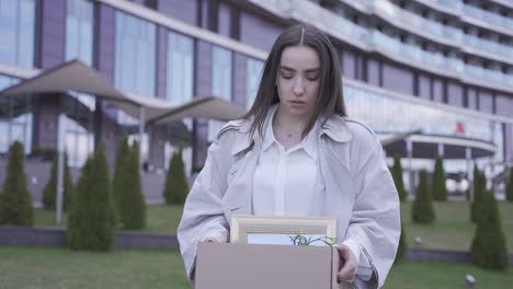 una mujer bonita camina frente a un edificio de oficinas. ella lleva sus cosas en una caja. ella ha sido despedida. ella está triste.