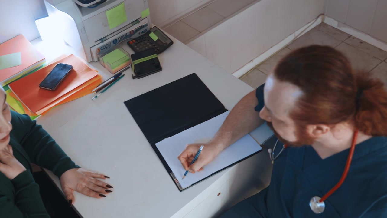 Paramédico realizando una consulta de salud a domicilio, mujer sentada en el escritorio. Clínico escribiendo notas en un portapapeles, estetoscopio visible, documentos y teléfono en la mesa. Diálogo de apoyo y ambiente tranquilo.