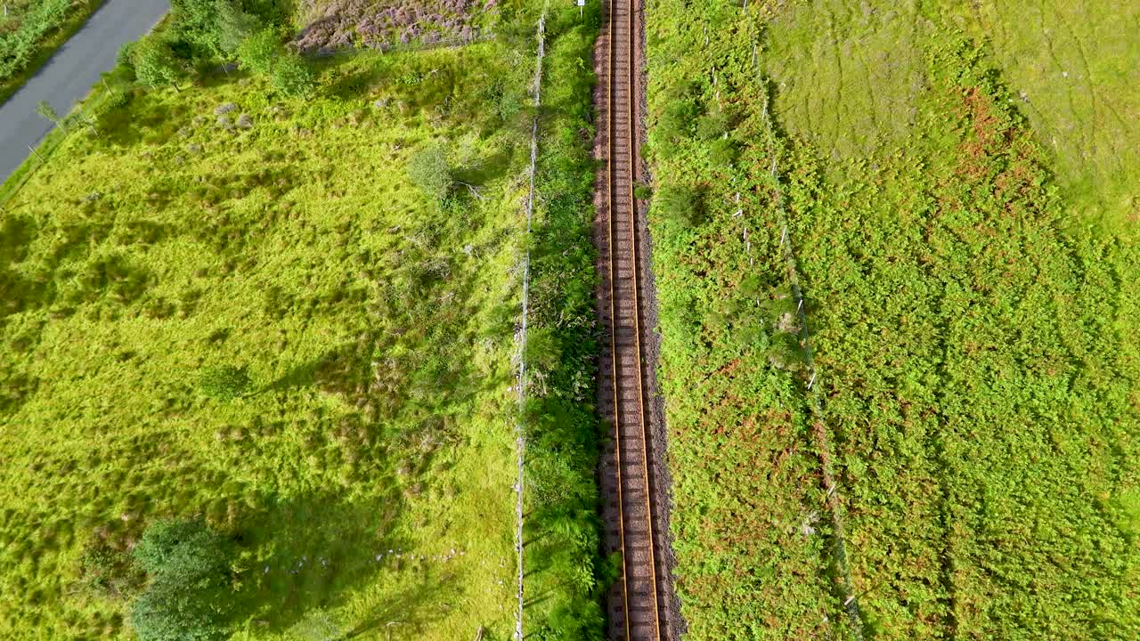 Drone camera moves above a single railway track running through lush green Highlands landscape, under bright daylight with clear, vibrant colors