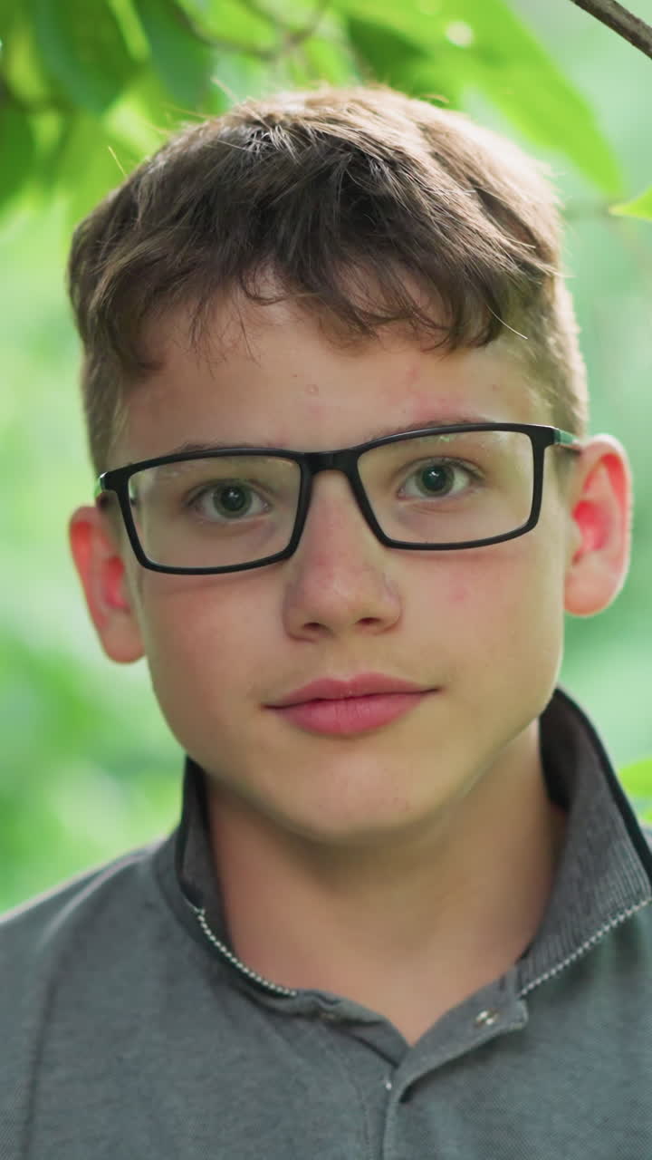 Close-up of young boy with pink lips wearing glasses and a grey top, blinking while focusing directly on the camera, a small insect flies in front of him, surrounded by blurred greenery