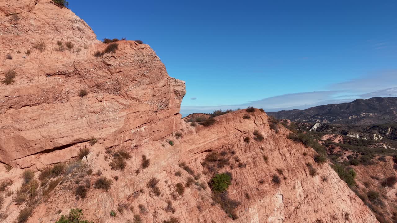 Cinematic aerial view of sunlit sandstone cliffs and rugged desert terrain with a distant mountain backdrop under a cloudless blue sky