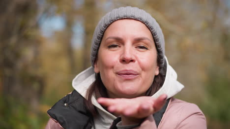 cheerful lady in ash beanie and pink jacket blowing dry leaves from her hand with a warm smile nails painted black, surrounded by blurred autumn trees