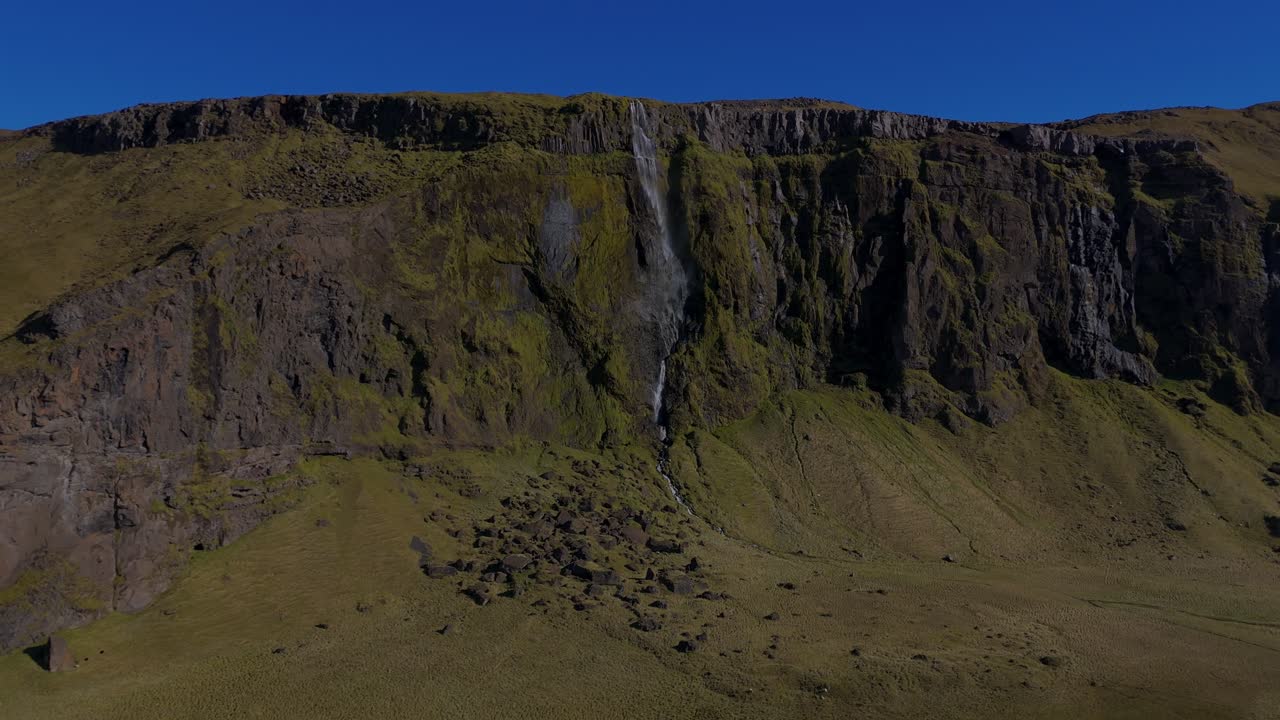 Ampla vista panorâmica de uma cachoeira alta e estreita caindo de um penhasco alto na Islândia. O terreno verde acidentado e as formações rochosas dramáticas destacam a escala e a beleza bruta da natureza nórdica intocada.