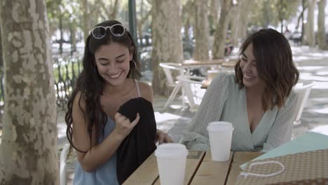 happy lady showing blouse to friend, smiling and drinking coffee