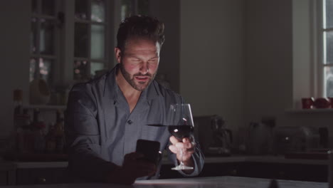 mature man wearing pyjamas sitting in kitchen with glass of wine at night using mobile phone