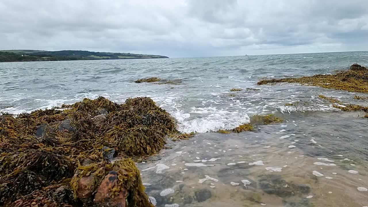 calma las ondas del océano en cámara lenta lavando a través de las rocas de la isla de anglesey en la playa de arena, mañana nublada