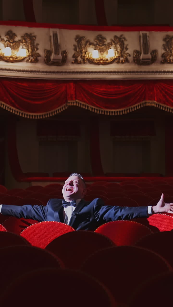 Successful man in suit alone in large auditorium. Actor man is sitting among empty red armchairs and singing in opera house. Vertical video