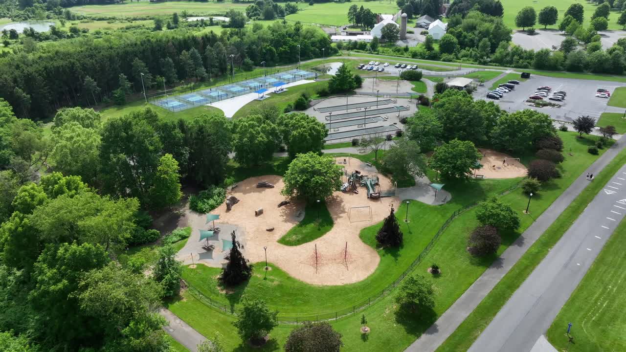 Aerial flyover empty american public park with playground, skatepark, parking area of cars, tennis courts and green trees on summer day