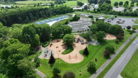 aerial flyover empty american public park with playground, skatepark, parking area of cars, tennis courts and green trees on summer day