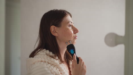 woman does microcurrent procedure for face lifting. brunette woman in robe makes massage movements on face using microcurrent in spacious shower cabin