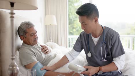 diverse male doctor testing blood pressure of senior male patient in bed at home, slow motion