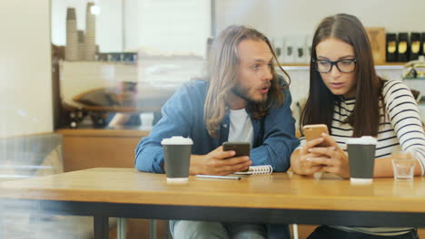 caucasian man and woman friends using smartphone and talking while they are sitting at a table in a cafe