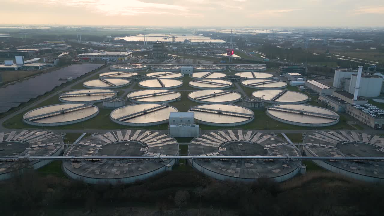 Kinoreife Drohnenaufnahme, die symmetrische kreisförmige Absetzbecken in einer modernen Kläranlage bei Sonnenaufgang zeigt, mit spiegelnden Wasseroberflächen und industrieller Infrastruktur.