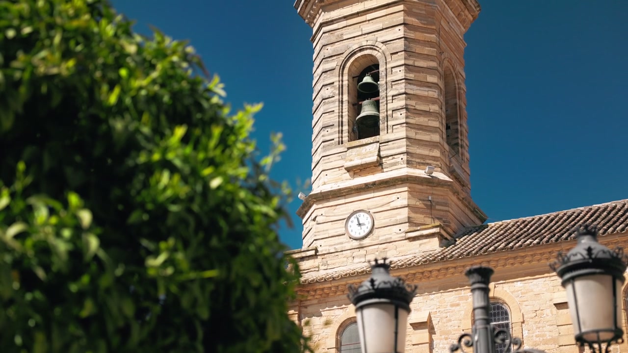 Un teleobiettivo inquadra dal basso verso l'alto una chiesa a Porcuna, in Spagna, mostrando la facciata in pietra e la torre dell'orologio con fogliame verde e lampioni decorati in primo piano