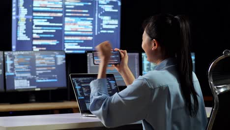 back view of asian female programmer looking at smartphone then celebrating the success while writing code by a laptop using multiple monitors showing data on desktops in the office