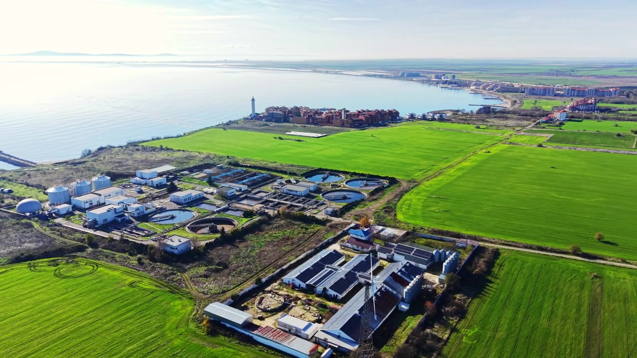 Aerial view of a wastewater plant beside the sea, Bulgaria. Elevated drone shot of treatment tanks, green fields, coastal town and lighthouse on the horizon.