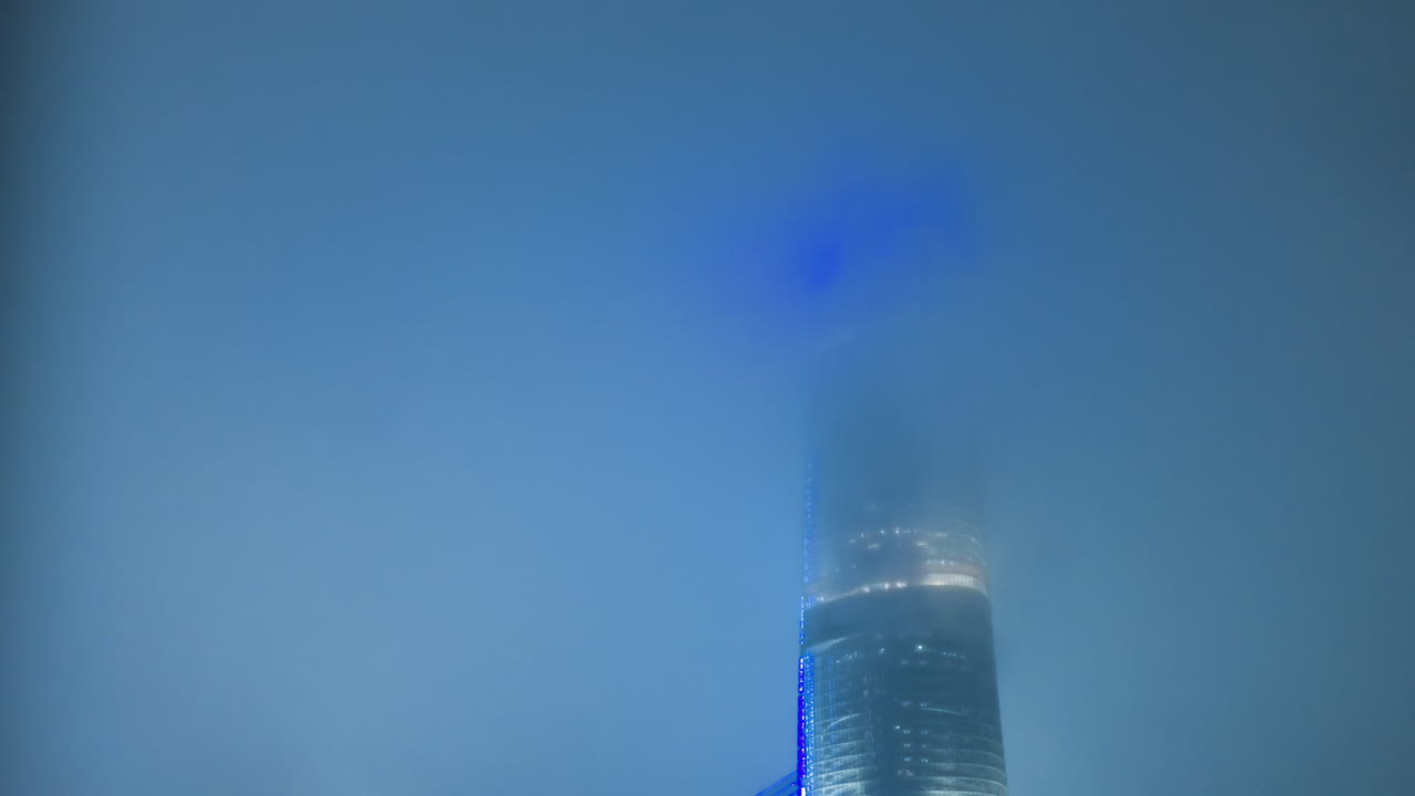 Blue tower shrouded in fog with glowing beacon and red navigation lights piercing mist over quiet metropolis at night, cinematic atmosphere with soft blur, cool blue palette, reflection on glass
