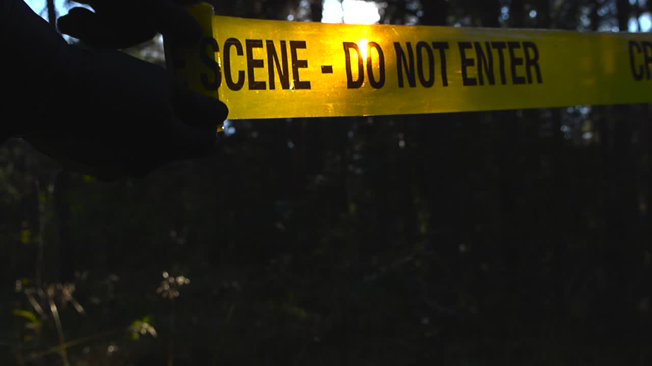 Police detective or investigator unrolling a yellow colored Crime Scene forensic investigation tape in front of a dark bokeh blurry forest with sunshine backlighting the hands and the ribbon, slow-mo