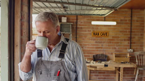 Portrait-Of-Mature-Male-Wearing-Overalls-In-Garage-Workshop-With-Hot-Drink