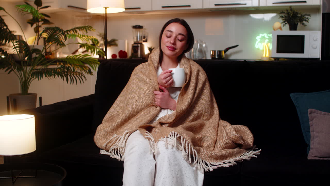 Giovane donna con coperta che tiene in mano una tazza di tè, sorseggia lentamente, mani calde, riposo tranquillo, serata intima e rilassante