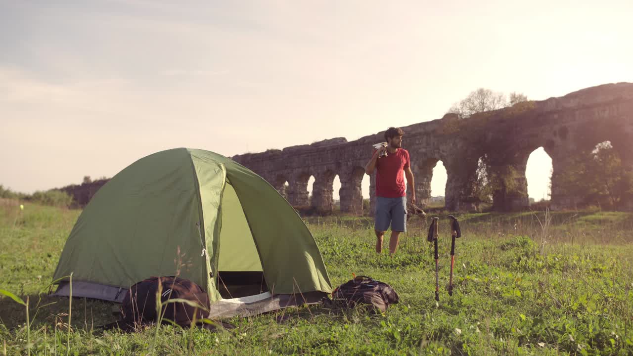 젊은 남자가 캠핑 텐트 밖에서 배을 들고 기타를 들고 로마의 수로 앞에서 풀 위를 습니다. parco degli acquedotti에 있는 아치 파크의 폐허는 해가 지는 동안 지상에서 느린 움직임을 유지합니다.