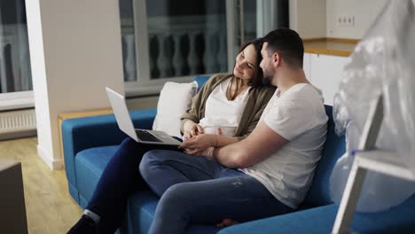 Lovely-pregnant-woman-and-a-positive-man-are-using-a-laptop-sitting-on-the-sofa-in-a-new-living-room