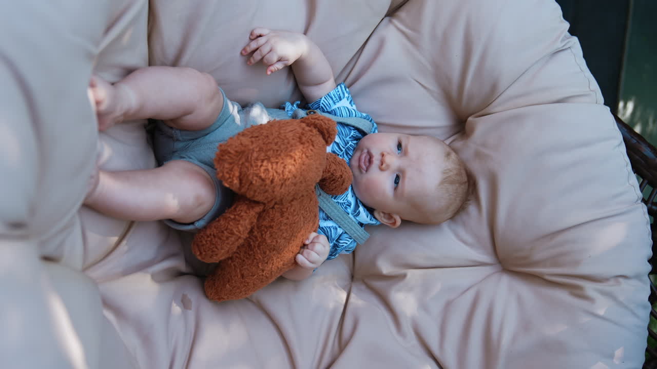 Adorable Caucasian infant lies in hanging chair with a teddy. Cute baby boy resting outdoors. Vertical screen.