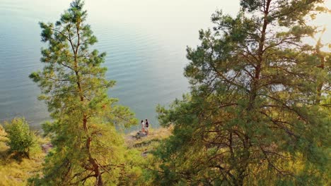 aerial view on the young couple is hugging near water. beautiful green pine trees on the water background and happy loving man and woman spending time together.