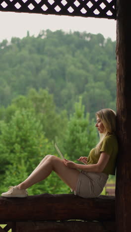 pretty blonde woman writer works on laptop computer sitting on terrace log railing at ecoresort at highland on overcast day side view slow motion