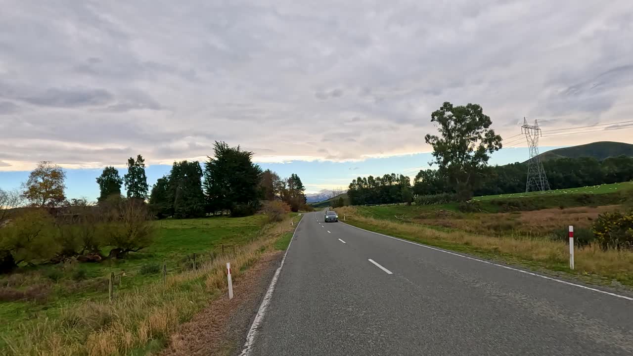 A serene drive along a rural highway in Akaroa, New Zealand, showcasing lush greenery, distant hills, and a passing vehicle