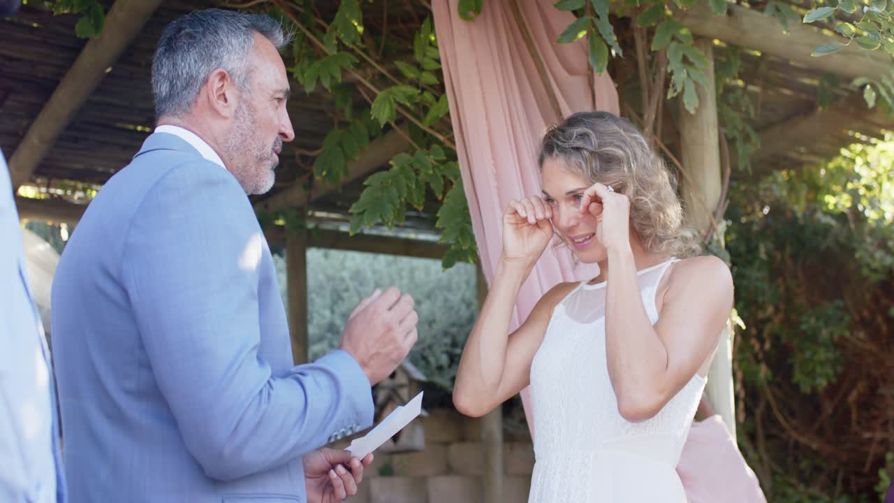 Man in light blue suit reading printed vows at wedding under wooden pergola, woman wiping tears