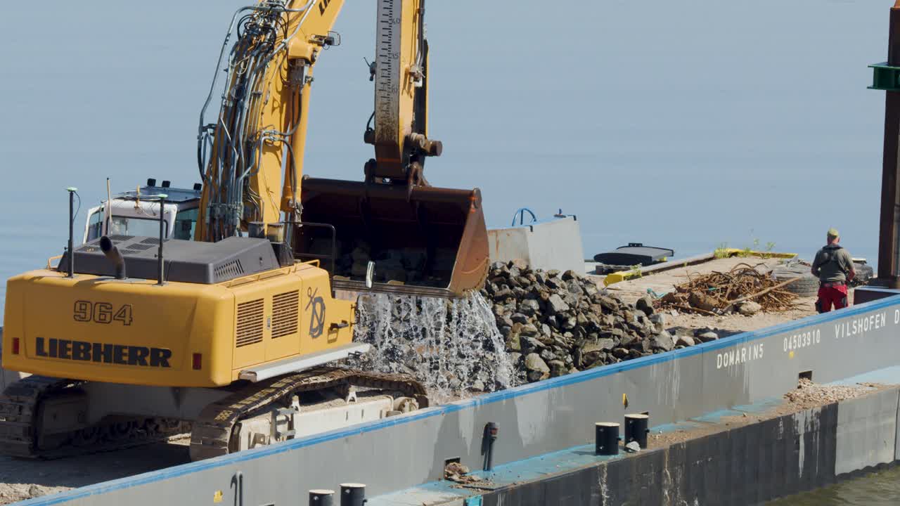 A large excavator deposits rocks onto a barge at a waterfront construction site in Dresden, Germany, under bright daylight with steady camera framing