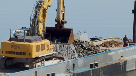 a large excavator deposits rocks onto a barge at a waterfront construction site in dresden, germany, under bright daylight with steady camera framing