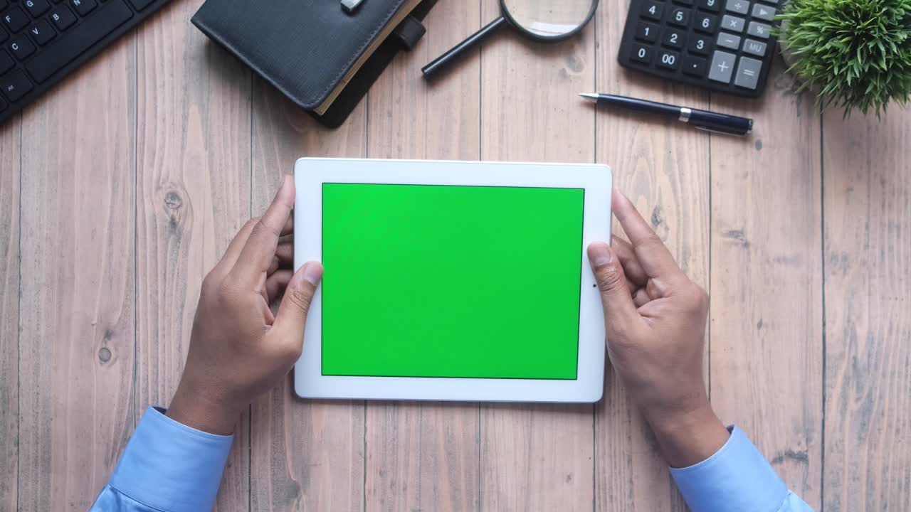 Person holding a tablet with a green screen on a wooden desk