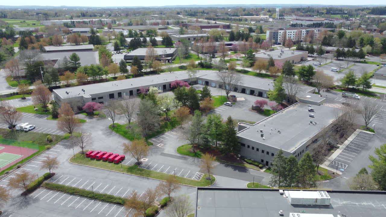 A rotating drone shot captures a tree-lined commercial park in Lancaster, PA. Blossoming spring trees surround low-rise office buildings and quiet parking lots, creating a peaceful business scene.