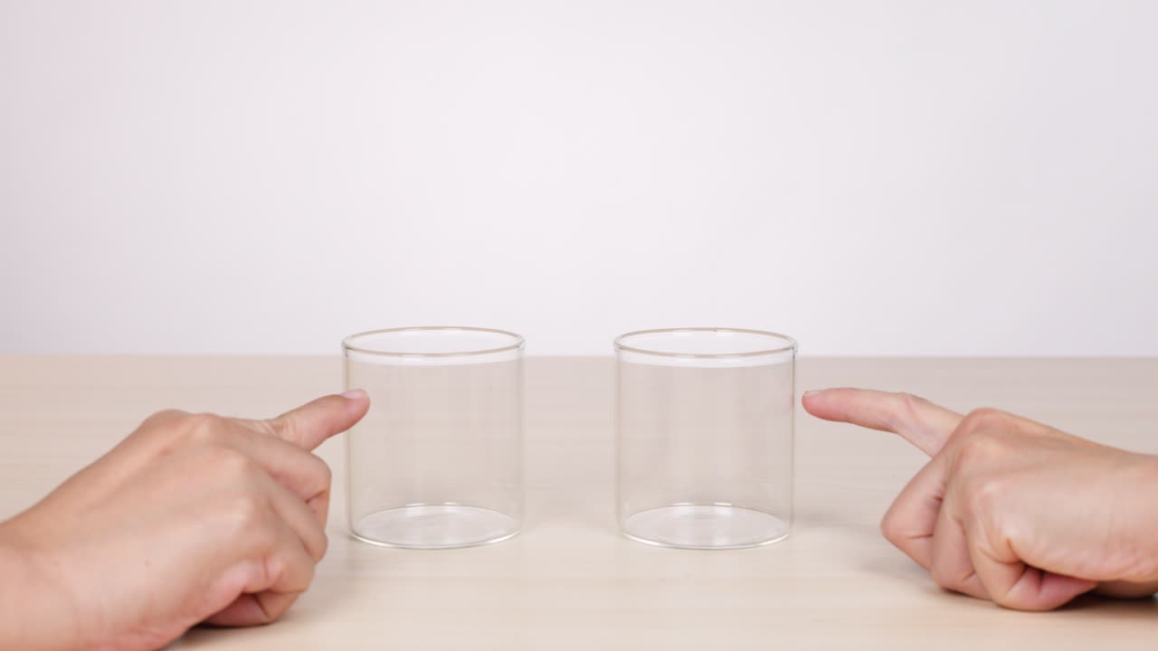 Two hands point at empty glasses on a table, neutral lighting, static camera, minimal movement