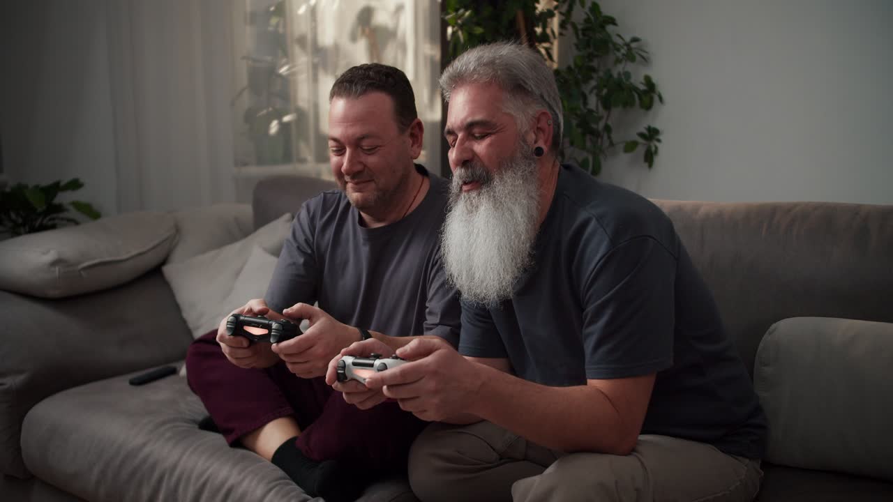 un hombre feliz con cabello gris una barba exuberante y un pendiente en la oreja junto con su novio una morena de mediana edad con paja en una camiseta gris jugando videojuegos usando joysticks mientras está sentado en el sofá en un apartamento moderno por la noche