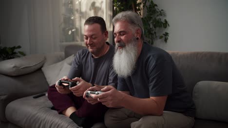 a happy man with gray hair a lush beard and an earring in his ear together with his boyfriend a middle-aged brunette with stubble in a gray t-shirt play video games using joysticks while sitting on the sofa in a modern apartment in the evening