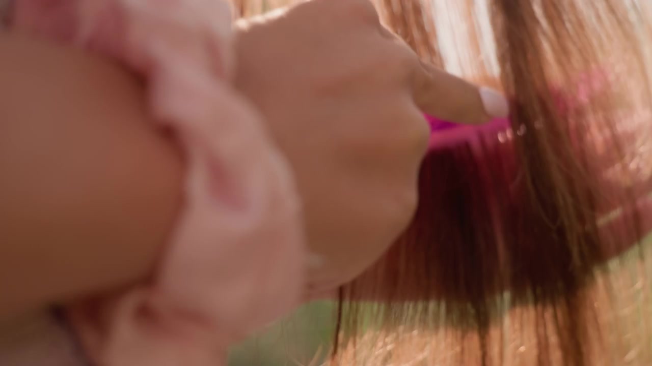Primer plano de una mujer joven con cabello largo castaño claro, que toca suavemente y pasa sus dedos por los mechones. El enfoque suave resalta los detalles de la textura, mientras un accesorio de tela rosa