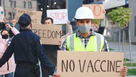 caucasian builder wearing protective mask and helmet holding no vaccine" singboard and looking at the camera in a protest against covid 19"