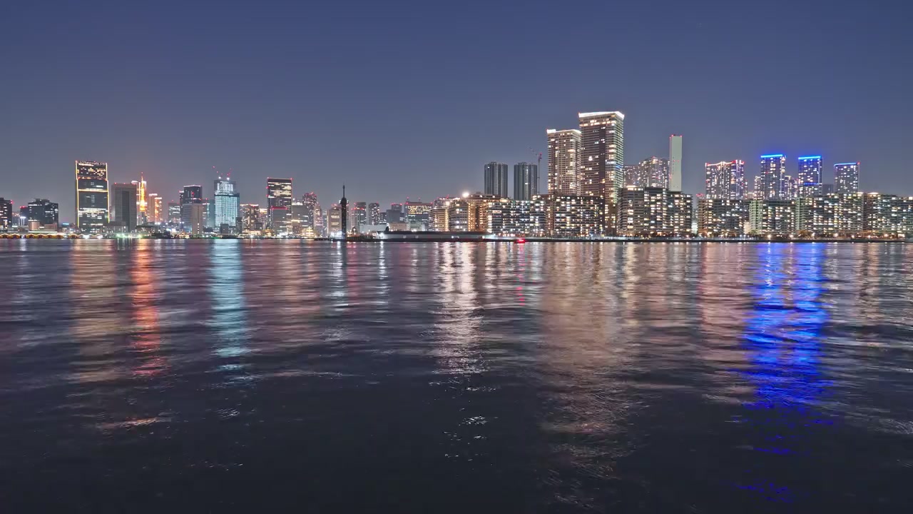 Wide static time-lapse of Tokyo Bay at night, dense glittering residential and commercial towers spanning the full horizon, multicolored light reflections shimmering on gently rippling dark water.