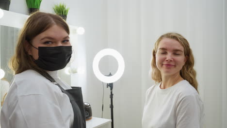 client and technician share warm smile while facing each other in beauty salon with ring light, vanity mirror, and modern white curtain before shifting attention to camera in relaxed moment