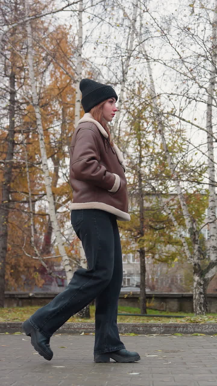 Side view of lady in black knit cap, brown shearling jacket, and black trouser walking peacefully through paved park on cold overcast day, light snowfall falling with bare trees and soft autumn backdrop