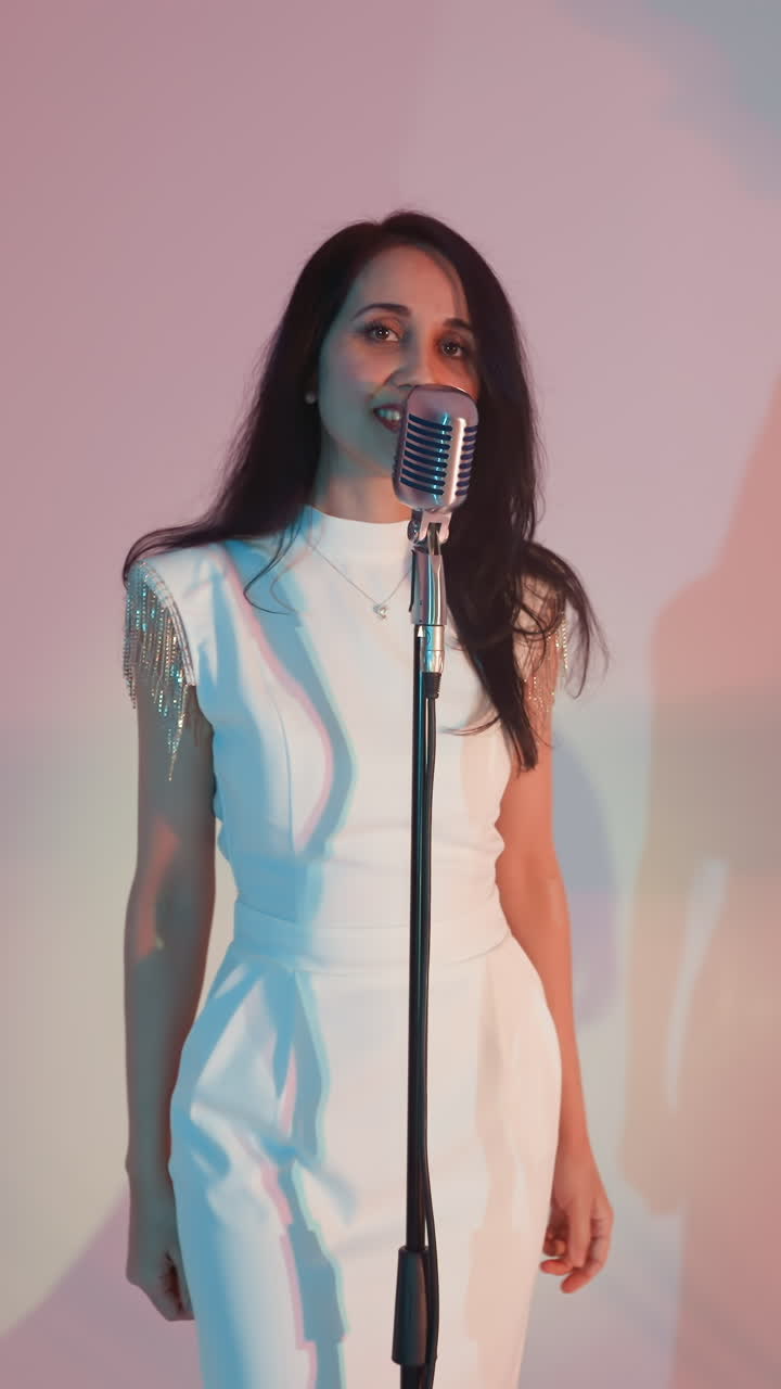 una vocalista con una túnica blanca canta apasionadamente mientras mueve sus manos. una guitarrista y una tecladista, ambas con trajes negros, tocan contra un fondo colorido. sombras en la pared