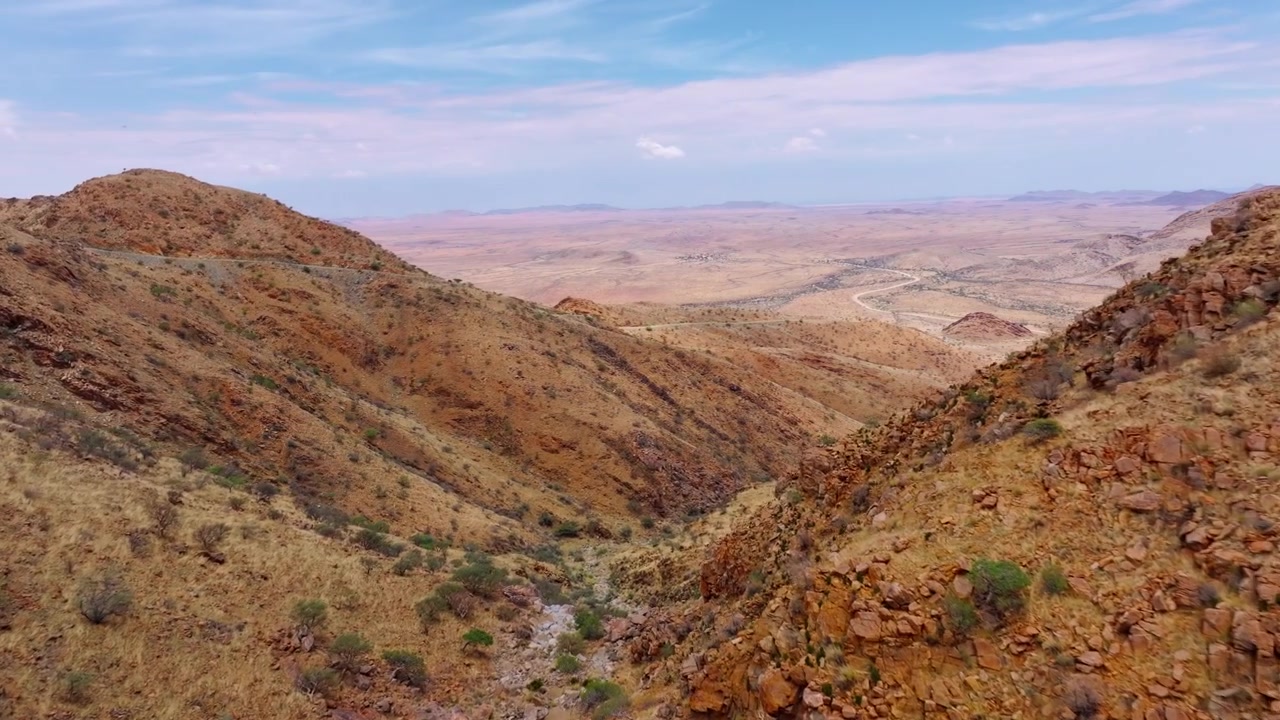 Drone view of layered arid mountain ridges under a partly cloudy sky in the Namib desert. A lonely gravel road snakes through the dramatic sun-baked mountains of the great escarpment.