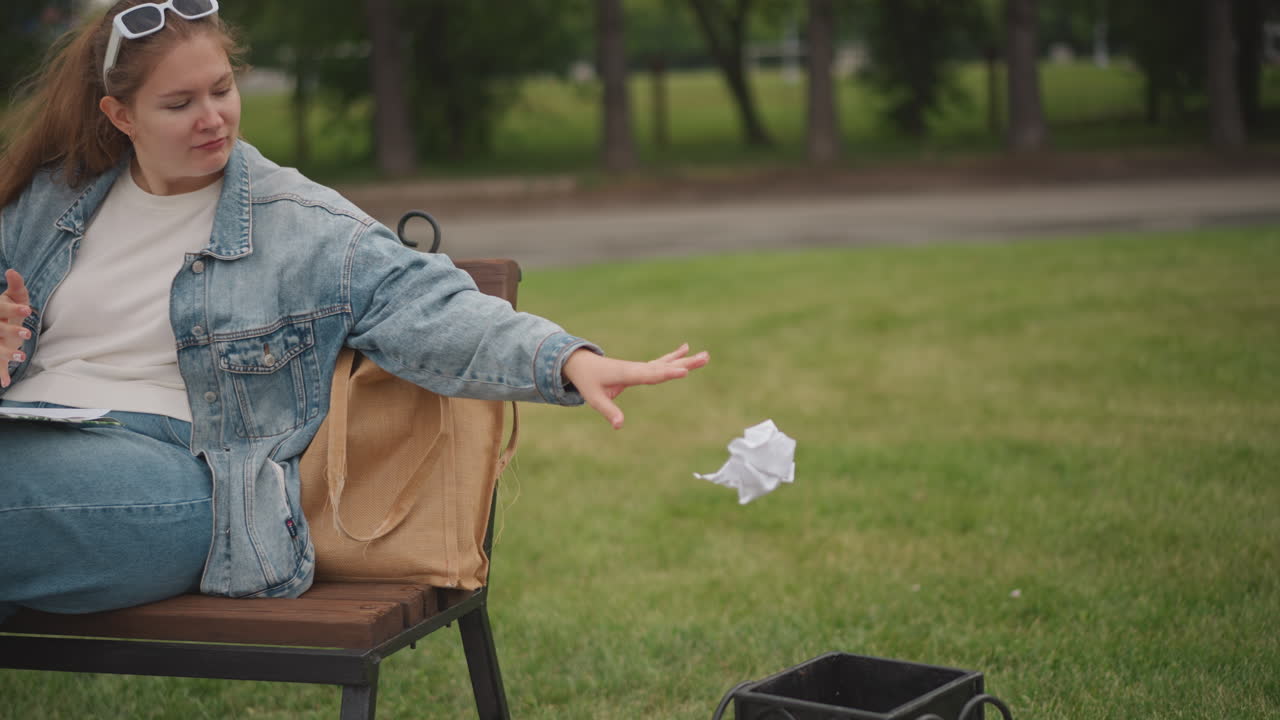 Una donna getta della carta nel cestino. Una donna in jeans smaltisce la carta su un tranquillo sentiero del parco. Una donna caucasica su una panchina del parco getta la carta nel cestino sorridendo dolcemente all'ambiente sereno.