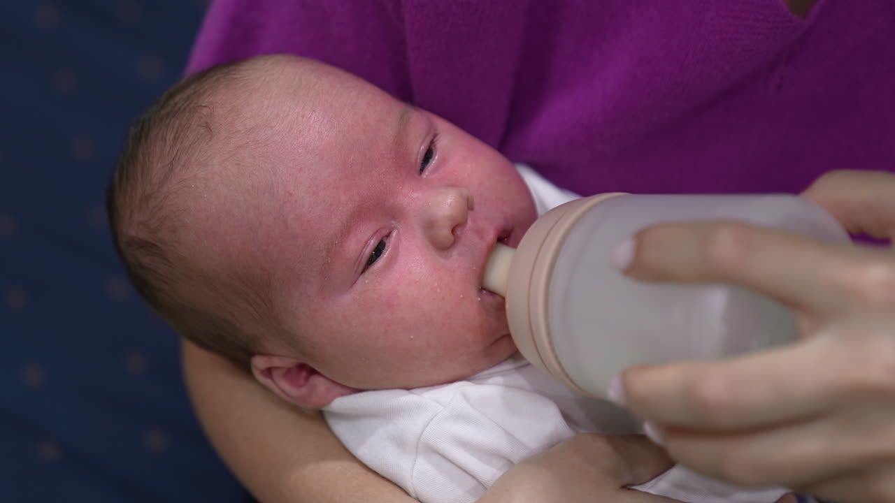 Adorable child in mother's hands ready to have meal. Little kid refuses to take the bottle first but then takes it and starts to eat. Close up.