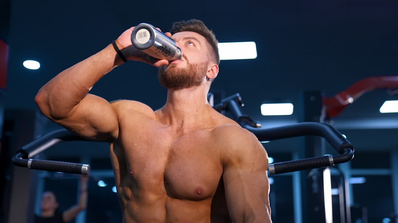 Man with perfect body train muscles on sport equipment and drinks water. View from the side on handsome man with perfect body.